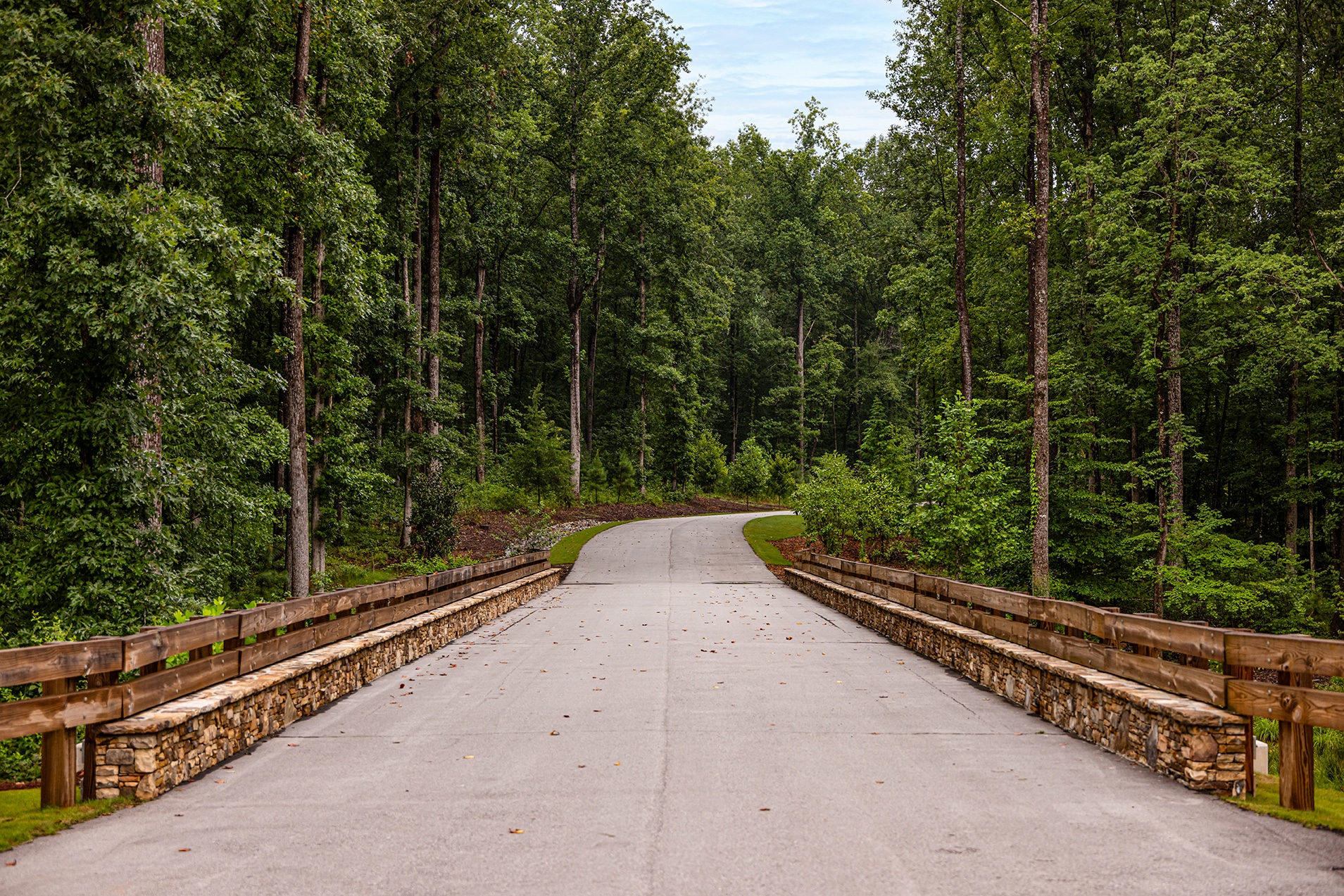 A two lane road leading over a bridge with stone sides, continuing into the woods